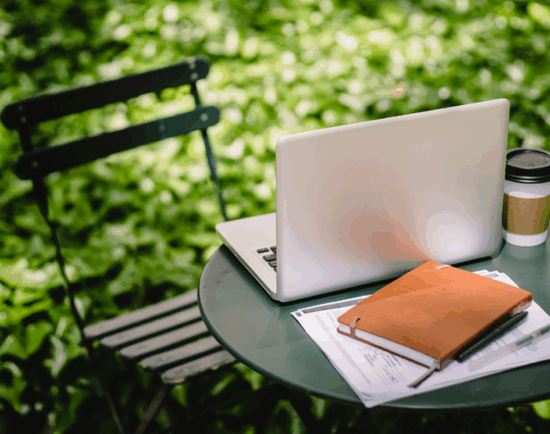 Laptop, journal, handouts, and mug on a small table in nature, symbolizing growth and sustainable leadership